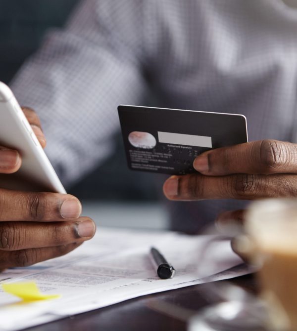 Cropped shot of African-American businessman paying with credit card online making orders via Internet. Successful black male holding plastic card making transaction using mobile banking application