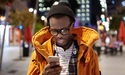 Headshot of handsome young Afro American student having walk around night city, holding mobile phone, using urban wifi, browsing pictures on social media. Modern technology and communication
