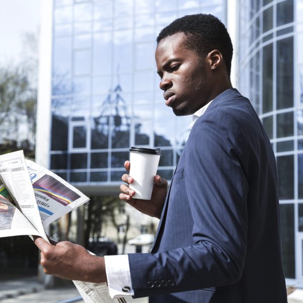 portrait-businessman-standing-front-building-holding-disposable-coffee-reading-newspaper